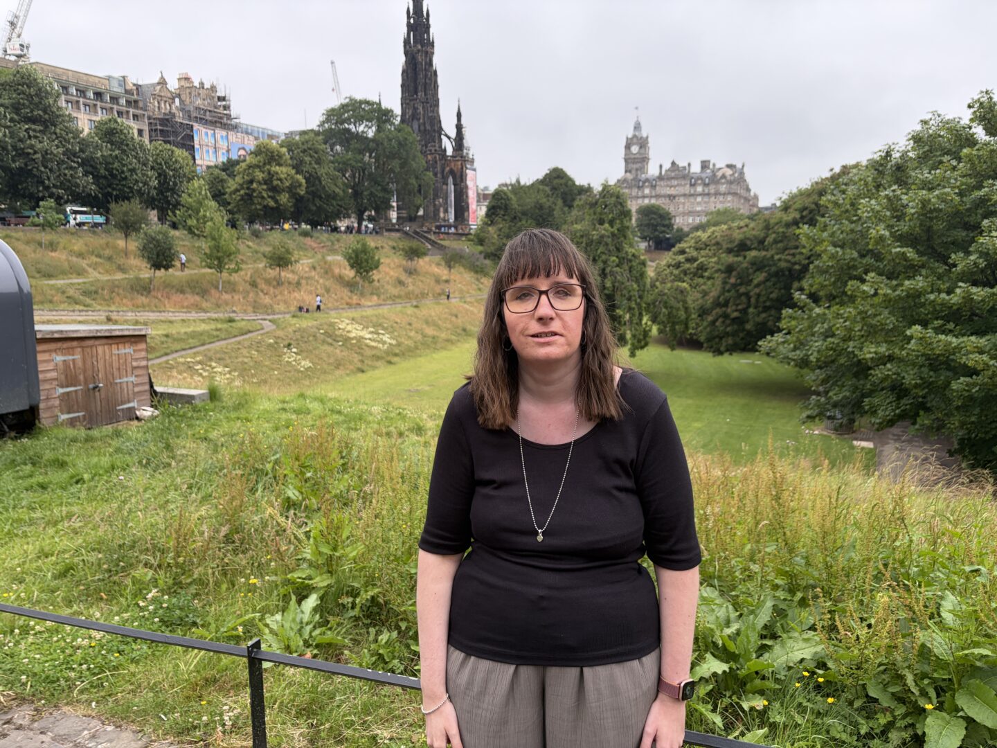 Holly stood in gardens with a tall building with a large tower and clock behind her, and a historic monument in the distance. She wears a black top, green trousers and a silver necklace.