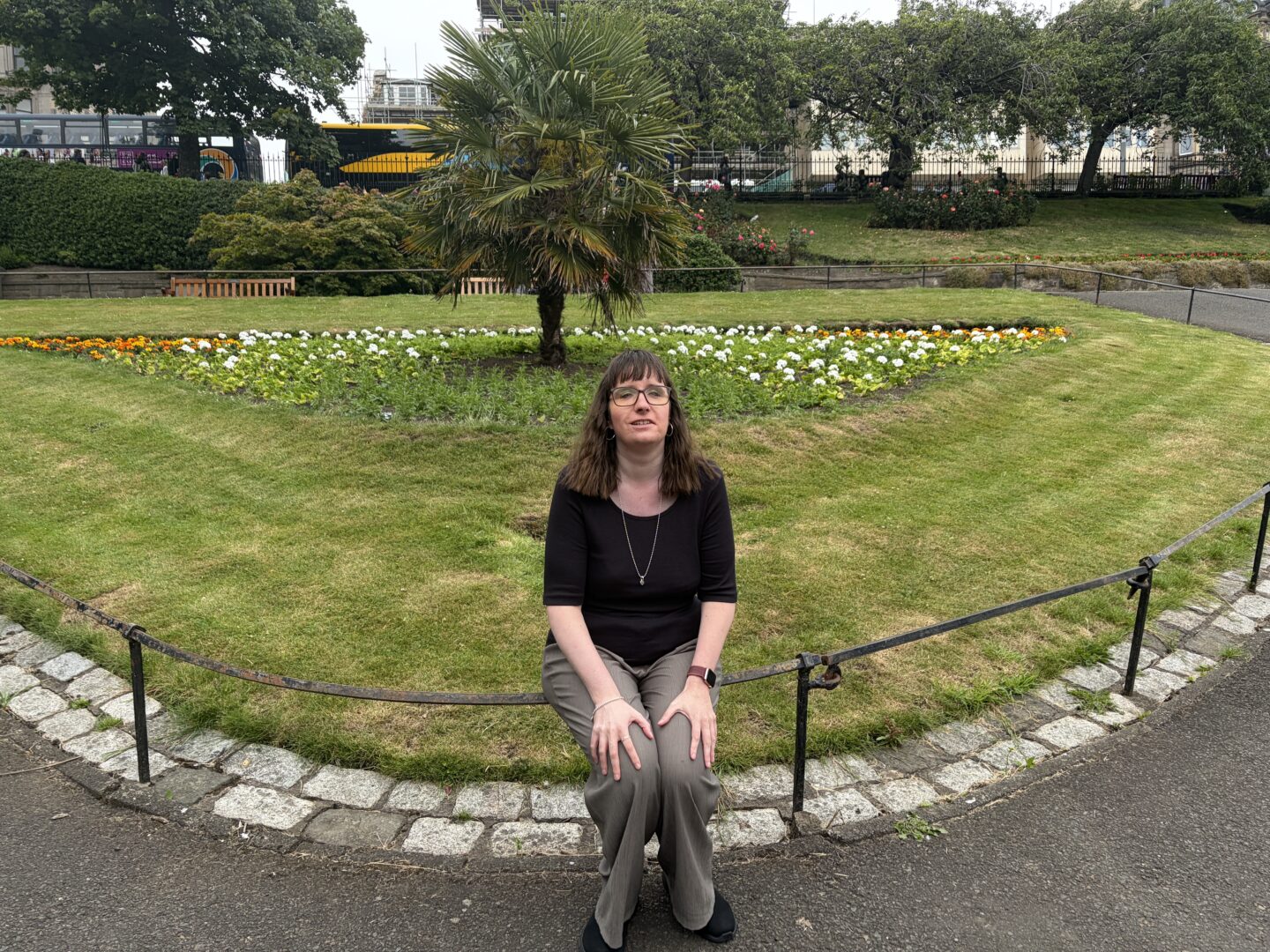 Holly sat on a metal railing in front of white flowers surrounding a large palm tree and a lawn. There are larger trees bordering a road in the background. She wears a black top, green trousers, black shoes and a silver necklace. She has her hands on her knees and is looking towards the camera.