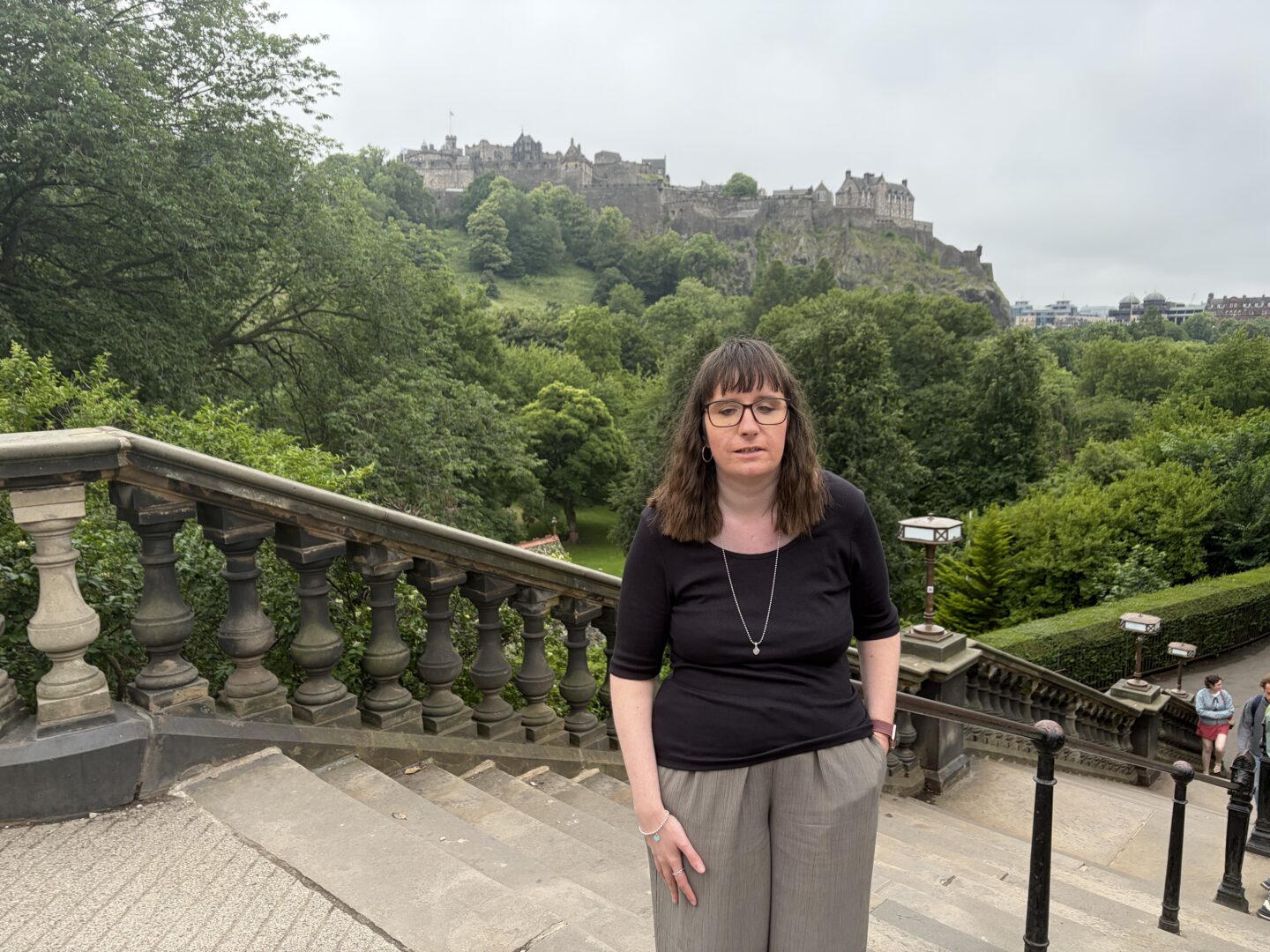 Holly stood at the top of stone steps with one hand in her pocket. There are bushes and green trees in the background, and Edinburgh Castle can be seen on the skyline in the distance. She wears glasses, a black top, green trousers and a silver necklace.