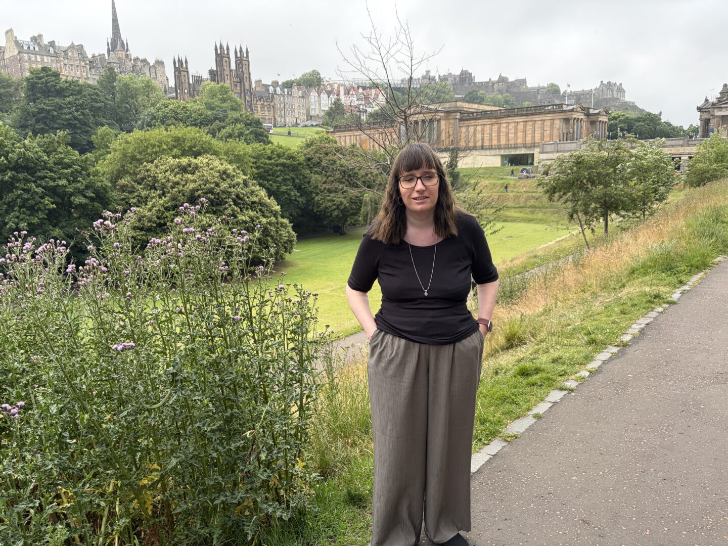 Holly stood on a paved path with her hands in her pockets. Grass and hedges are in the background. A large ornate building is in the foreground, with Edinburgh Castle in the distance. She wears glasses, a black three-quarter sleeved top, high waisted green trousers, black shoes and a silver necklace.