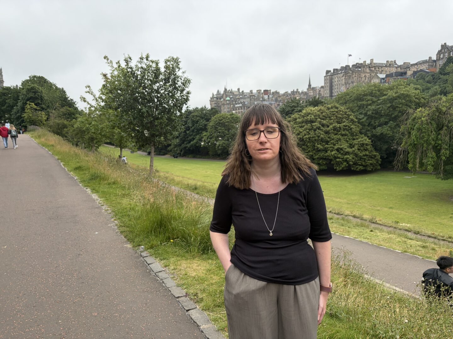 Holly stood on a path with one hand in her pocket. Hedges and trees are behind her. The historic Edinburgh Castle can be seen in the background. She wears glasses, a black three-quarter sleeved top, green trousers and a silver necklace.