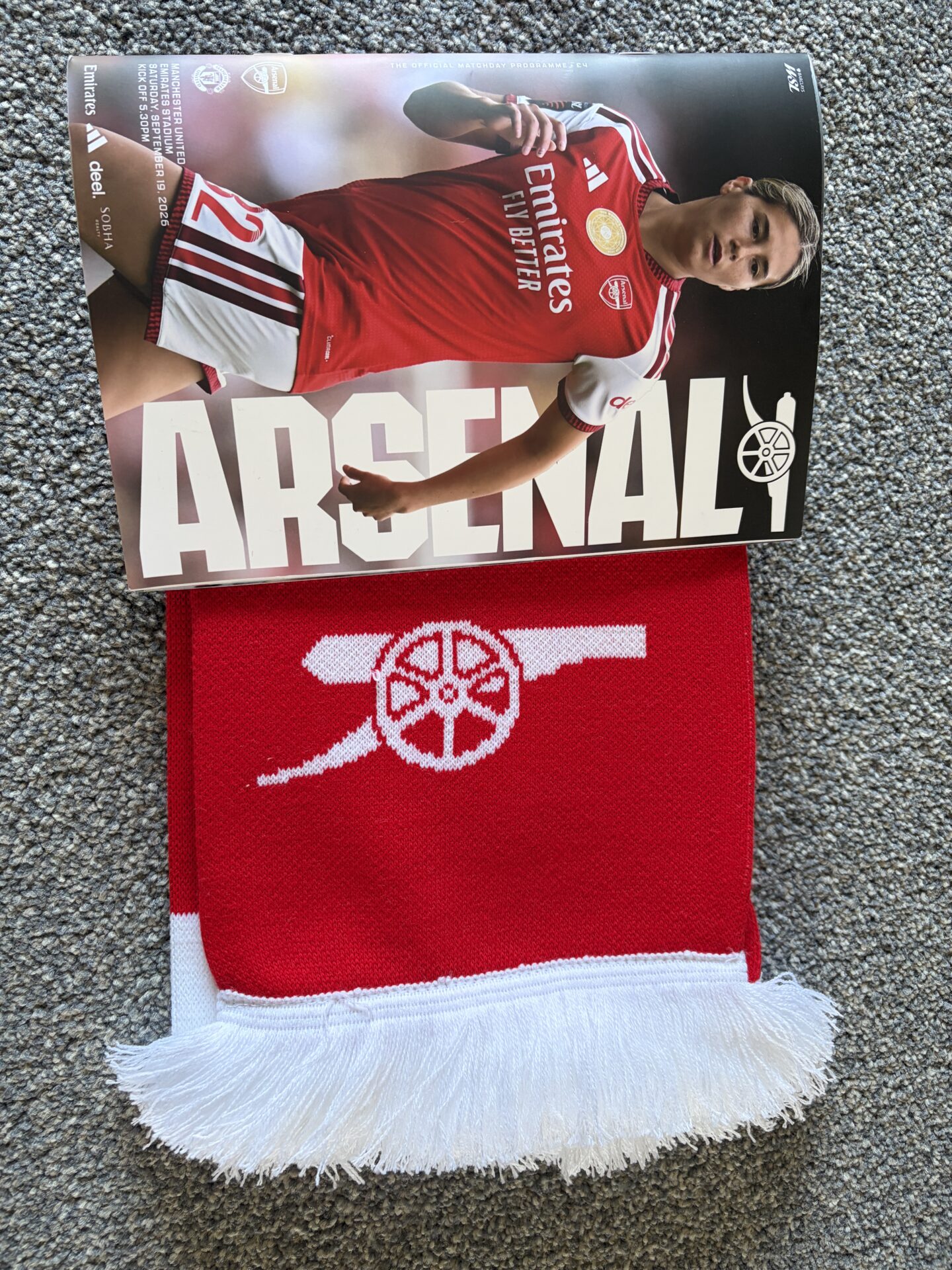 An Arsenal matchday programme placed on top of a red Arsenal scarf. The front cover of the programme shows an Arsenal player in the red and white home kit with white shorts and red socks. Across the bottom half of the cover, in large off white block letters, it says ‘Arsenal’ with the club’s cannon logo to the right. Smaller white text at the top left reads ‘The official matchday programme’ along with details of the fixture, date, and kick-off time. There are also sponsor logos including Emirates.