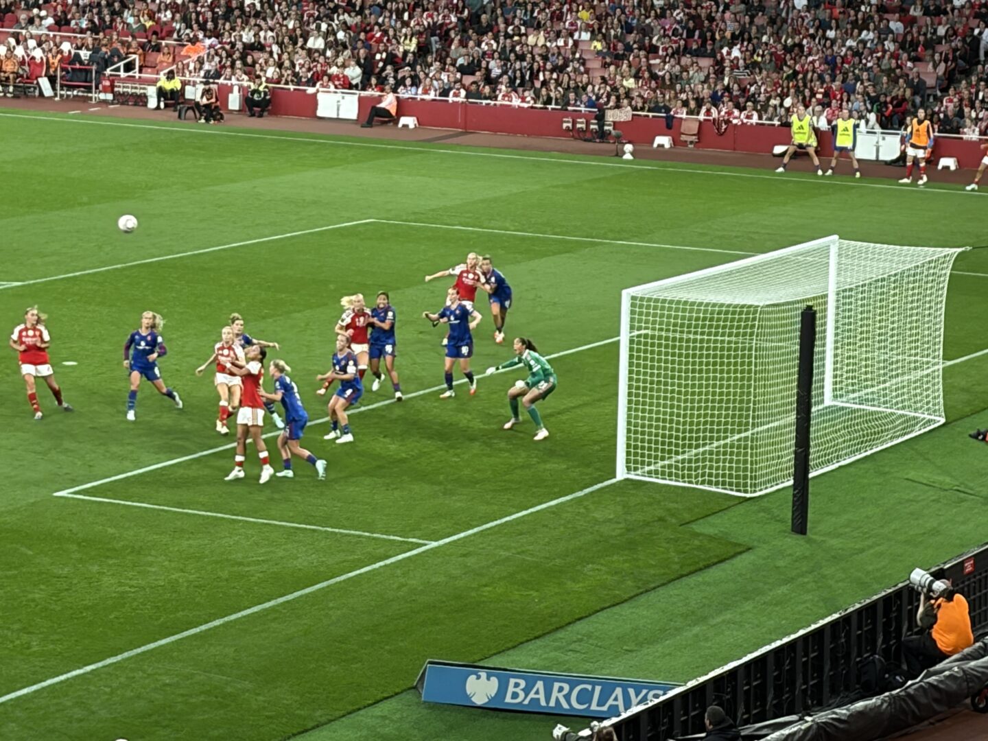 Arsenal players taking a corner, with Manchester United players defending. A white goal with netting is on the right side, and a goalkeeper in a green kit is slightly crouched near the goal line, ready to react. Arsenal are in their red home shirts, white shorts, and red socks; Manchester United are in their all blue away kit. Along the far touchline are substitutes and staff in fluorescent bibs standing near the dugout area, and a few security staff in yellow jackets. Fans pack the stands behind the pitch.