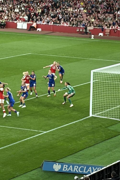 Arsenal players taking a corner, with Manchester United players defending. A white goal with netting is on the right side, and a goalkeeper in a green kit is slightly crouched near the goal line, ready to react. Arsenal are in their red home shirts, white shorts, and red socks; Manchester United are in their all blue away kit. Along the far touchline are substitutes and staff in fluorescent bibs standing near the dugout area, and a few security staff in yellow jackets. Fans pack the stands behind the pitch.