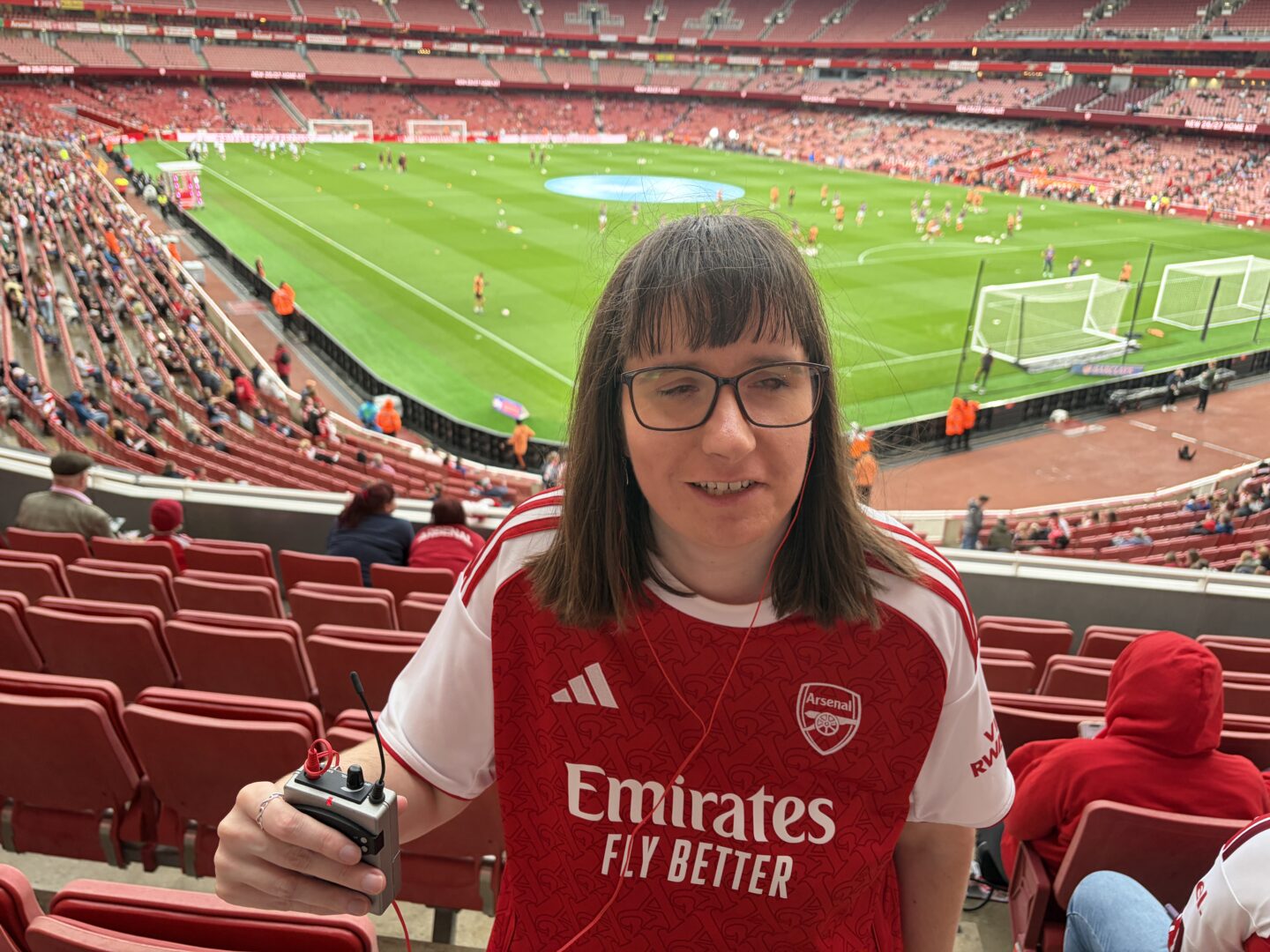 Holly stood in the stands at Emirates Stadium. She has a red earphone in one ear, and is holding her audio description receiver. There are rows of red seats around her. She wears a red and white Arsenal shirt with the club crest on the left chest and the sponsor text ‘Emirates Fly Better’ across the front. The pitch can be seen in the background. It has bright green grass and white goalposts at each end. Players and staff are on the pitch warming up before the match. Fans are seated around the stadium, and stewards in bright orange jackets stand near the edge of the pitch.