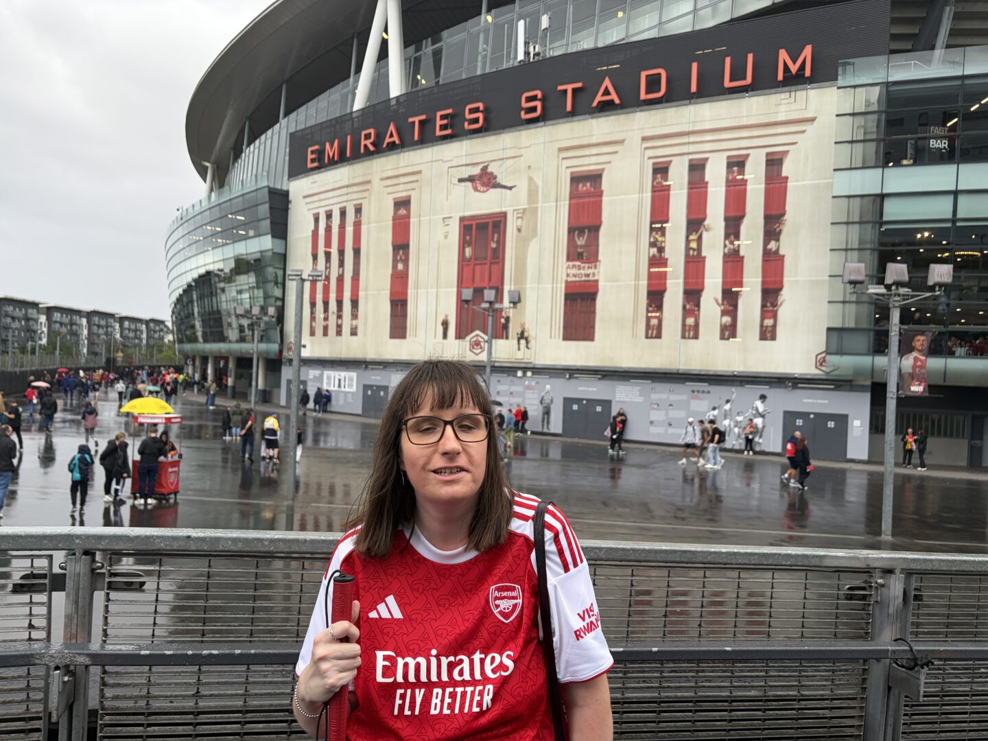 Holly stood in front of Emirates Stadium on a rainy day. She wears a red and white Arsenal shirt which features the Adidas logo, the Arsenal club crest, and the sponsor text ‘Emirates Fly Better’ on the front. The large exterior of Emirates Stadium is behind her. High above, big red letters spell out ‘Emirates Stadium’.