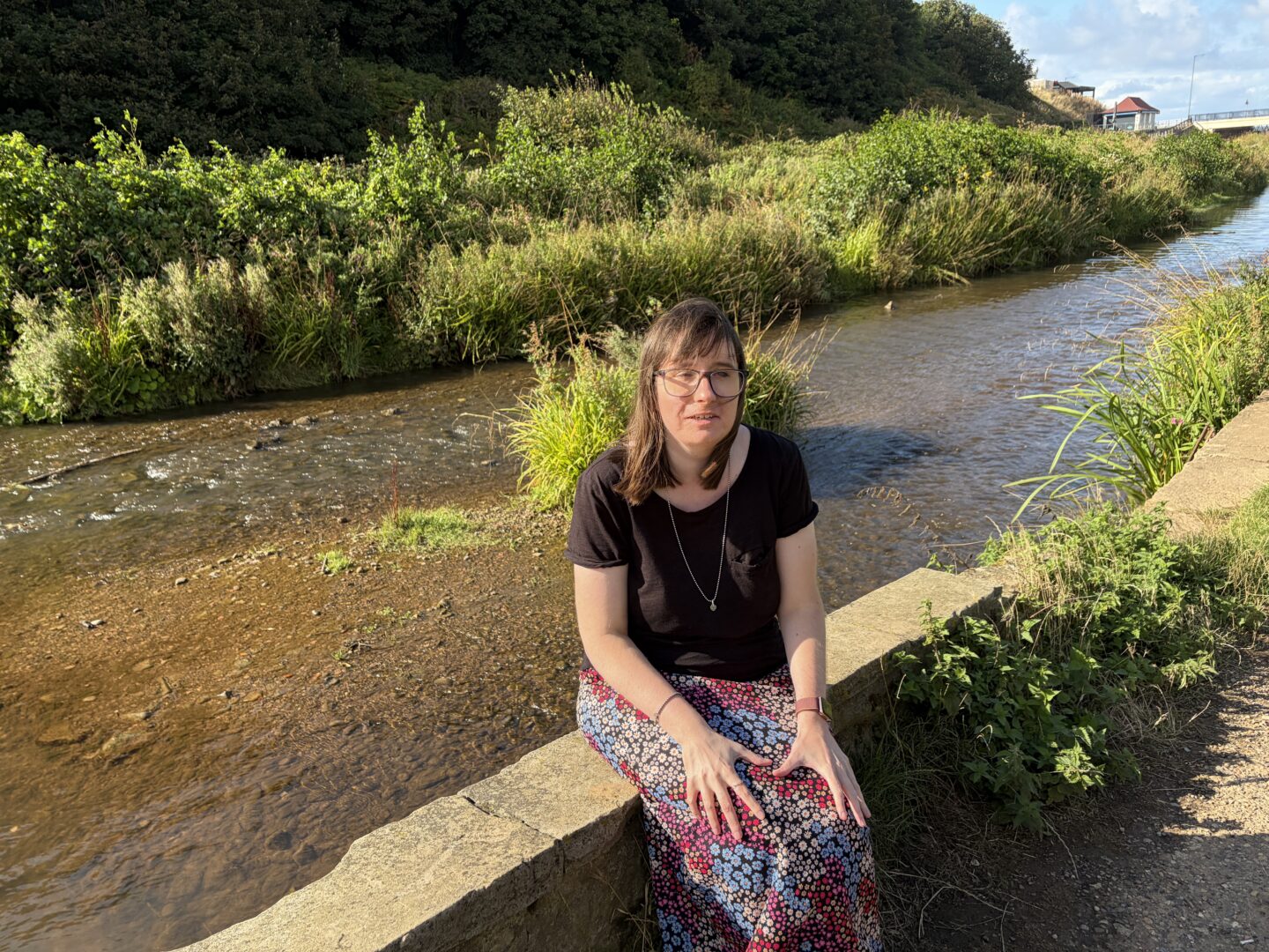 Holly sat on a low stone wall beside a shallow stream with a path running alongside it. Dense green bushes and trees line the far bank, forming a thick hedge along the water. There is a small bridge and a building in the background. She wears a black T-shirt, a long, multicoloured patterned floral skirt and a silver necklace.