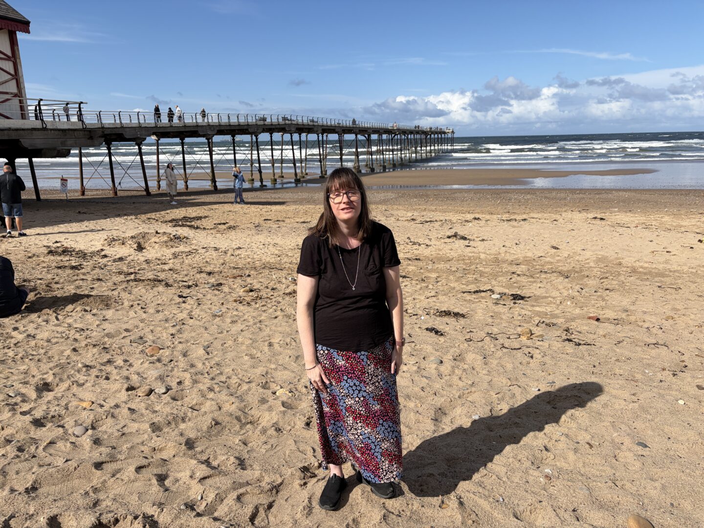 Holly stood on a beach with a long pier and the sea in the background. Waves can be seen breaking toward the shore in the distance. She wears a short sleeved black T-shirt, a colourful patterned skirt, black shoes, a silver necklace and glasses.