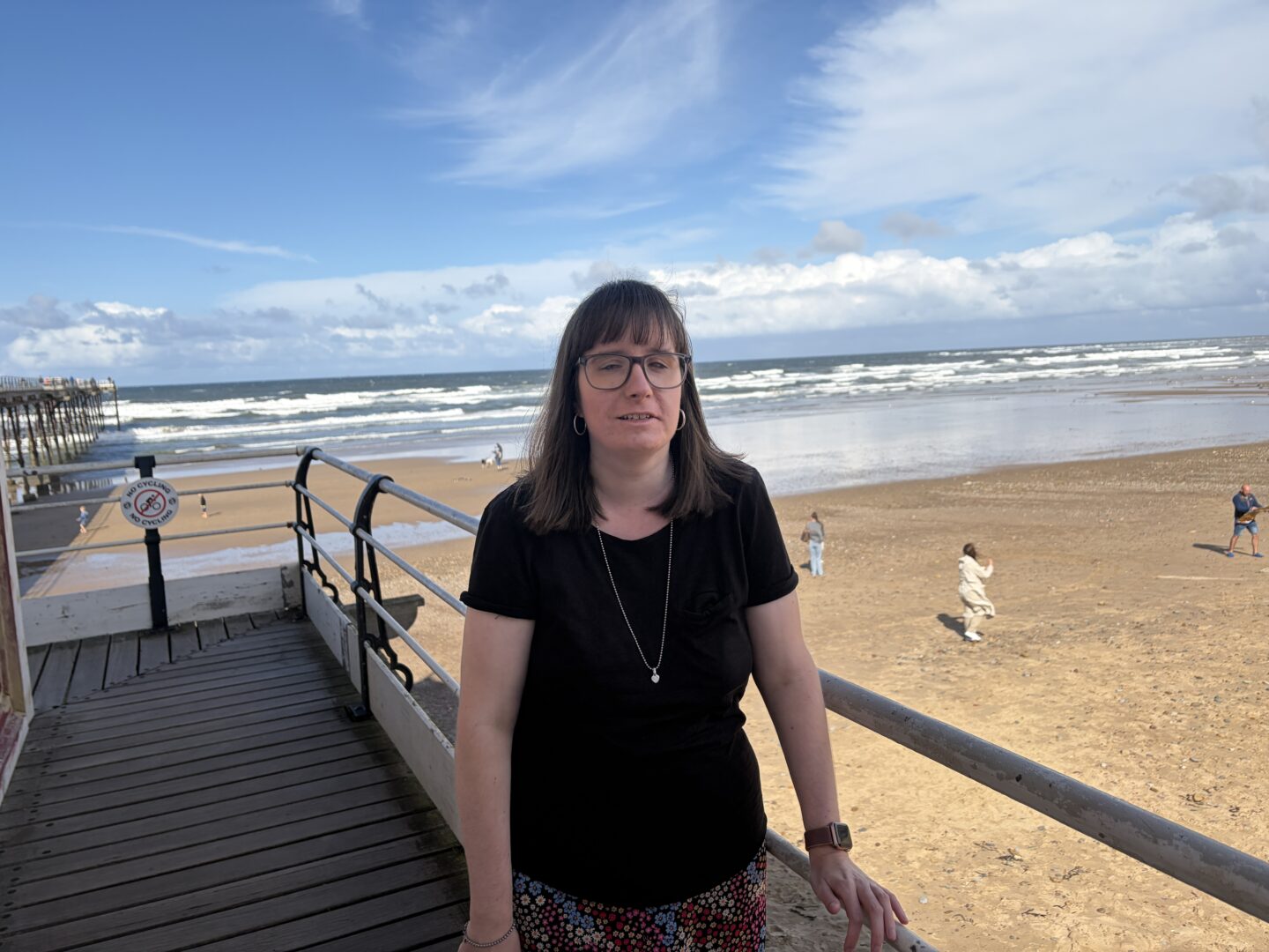 Holly stood on a pier, leaning against a railing by a sandy beach with the sea in the background. She wears a black T-shirt, a skirt with a colourful floral pattern, a silver necklace and glasses.