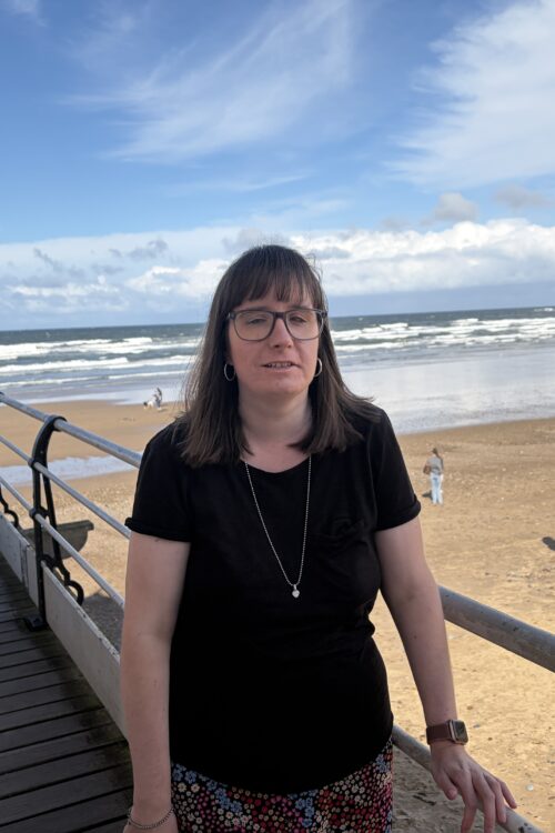 Holly stood on a pier, leaning against a railing by a sandy beach with the sea in the background. She wears a black T-shirt, a skirt with a colourful floral pattern, a silver necklace and glasses.