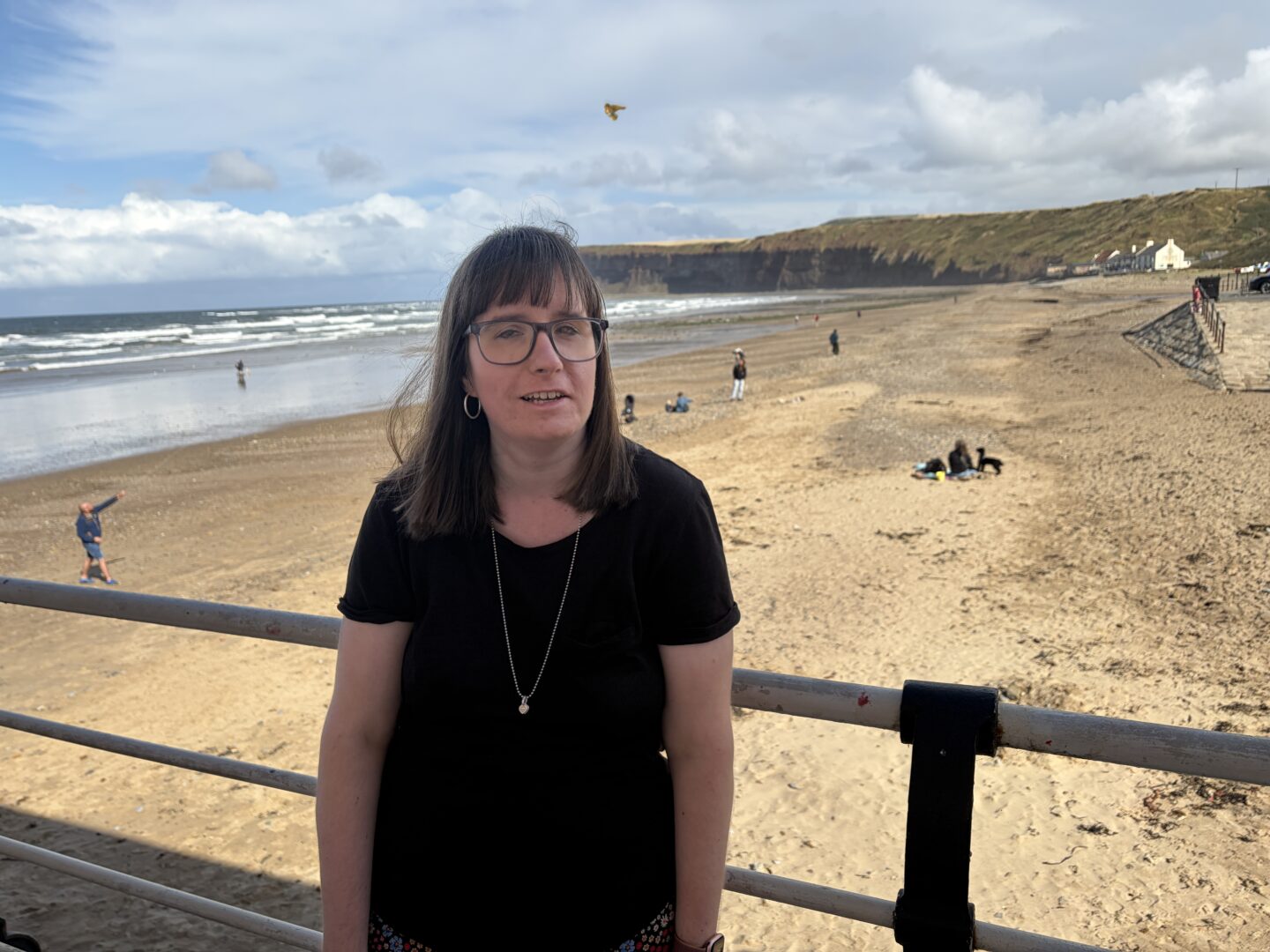 Holly stood on a seaside promenade in front of a sandy beach. The beach stretches into the distance towards low cliffs, with a line of small buildings visible. She wears a black short sleeved T-shirt, silver hoop earrings, a silver necklace and glasses, and is leant against a metal railing.