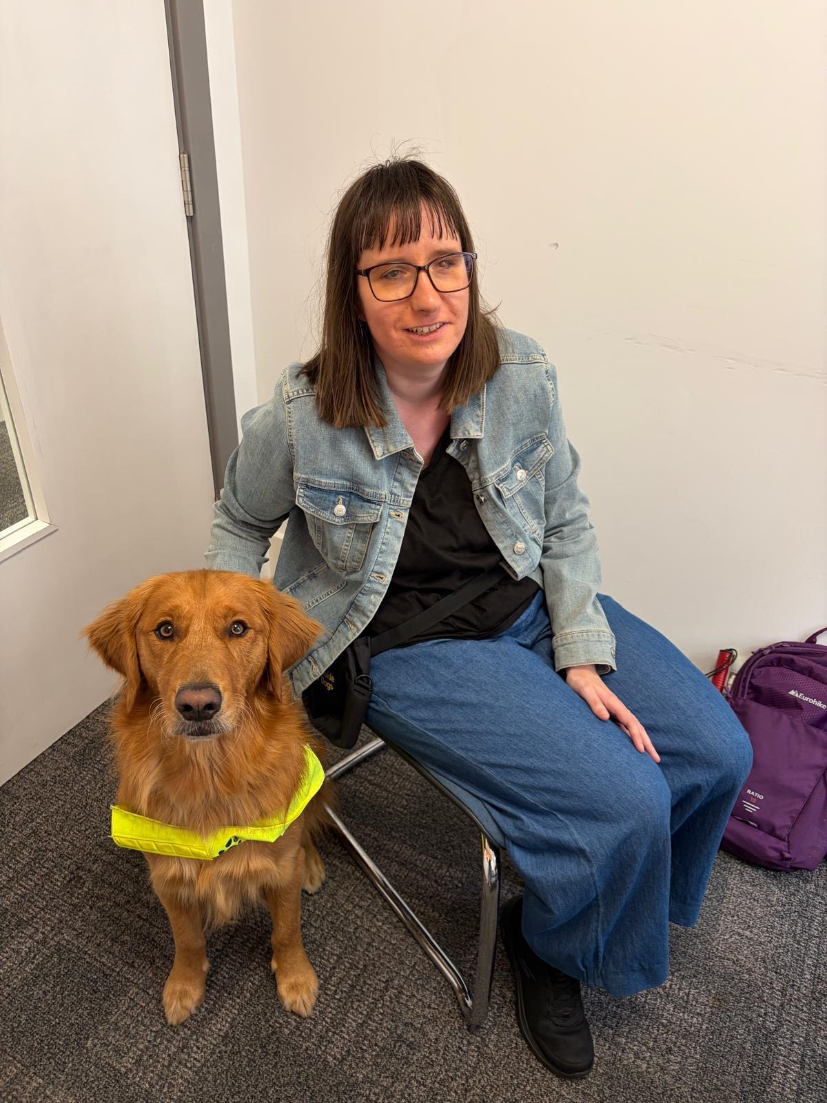 Holly sat on a chair next to Jukey, a golden retriever. She wears a blue denim jacket over a black top, blue jeans, black shoes and glasses, and is smiling. They’re both looking towards the camera.